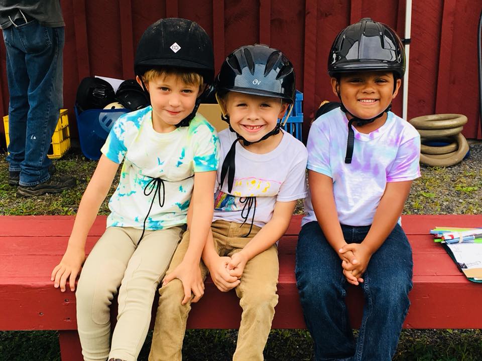 Three kids sitting on a red bench with helmets on smiling