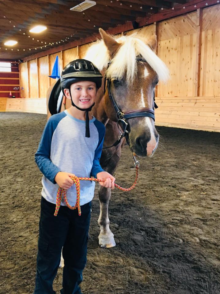 A boy smiling and wearing a helmet holding a lead rope with a horse on his right in an indoor ring at High & Mighty Therapeutic Riding and Driving Center