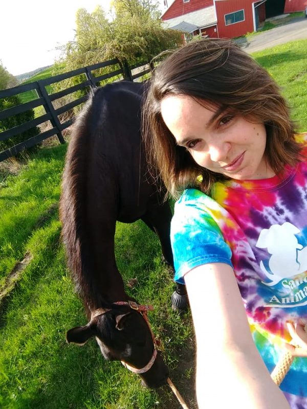 A selfie of a young woman wearing a tie dye shirt next to a dark horse in a grass field