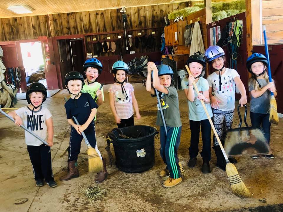 A group of young kids wearing helmets and smiling holding pitchforks and brooms with a muck tub inside the barn