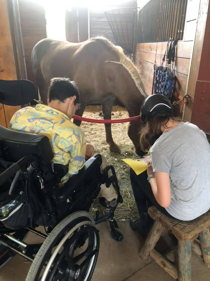 A boy in a wheelchair and a person sitting down in front of a horse's stall doing equine therapy