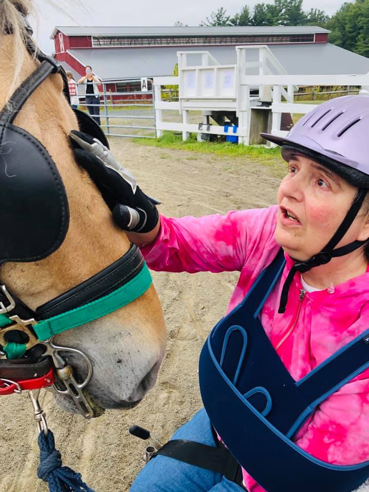 A woman in a wheelchair wearing a pink short and a helmet petting the forehead of a Fjord horse that is wearing blinders and a bridle