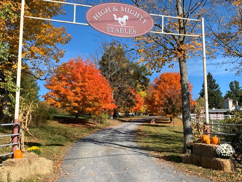 Farm entrance with an oval sign reading "High & Mighty Stables" and gravel driveway in the fall season