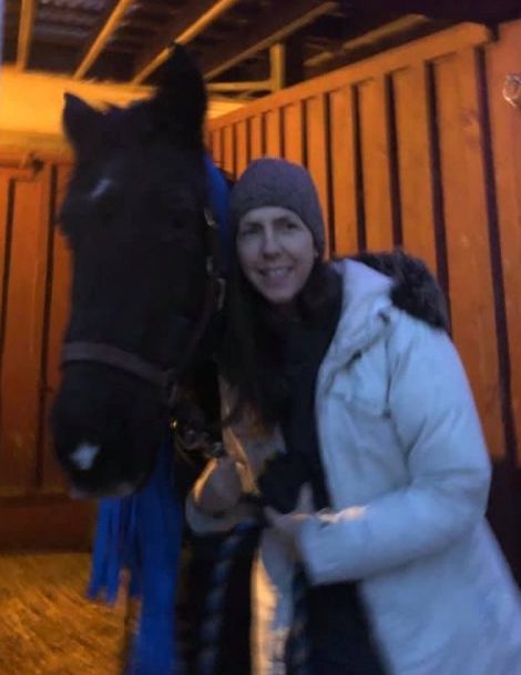 A woman in a white winter coat holding the lead rope of a dark brown horse and smiling in a horse's stall