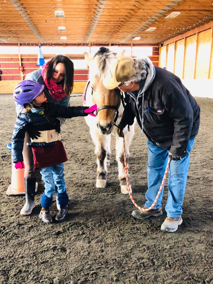 A woman holds a girl up while she pets a tan horse and a man stands to the right in the indoor arena at High & Mighty Therapeutic Riding and Driving Center