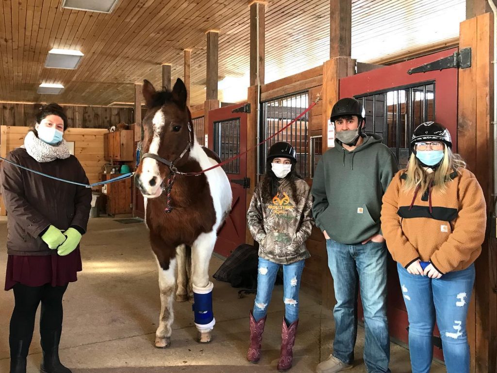 Four people standing next to a brown and white paint horse on the crossties in the barn at High & Mighty