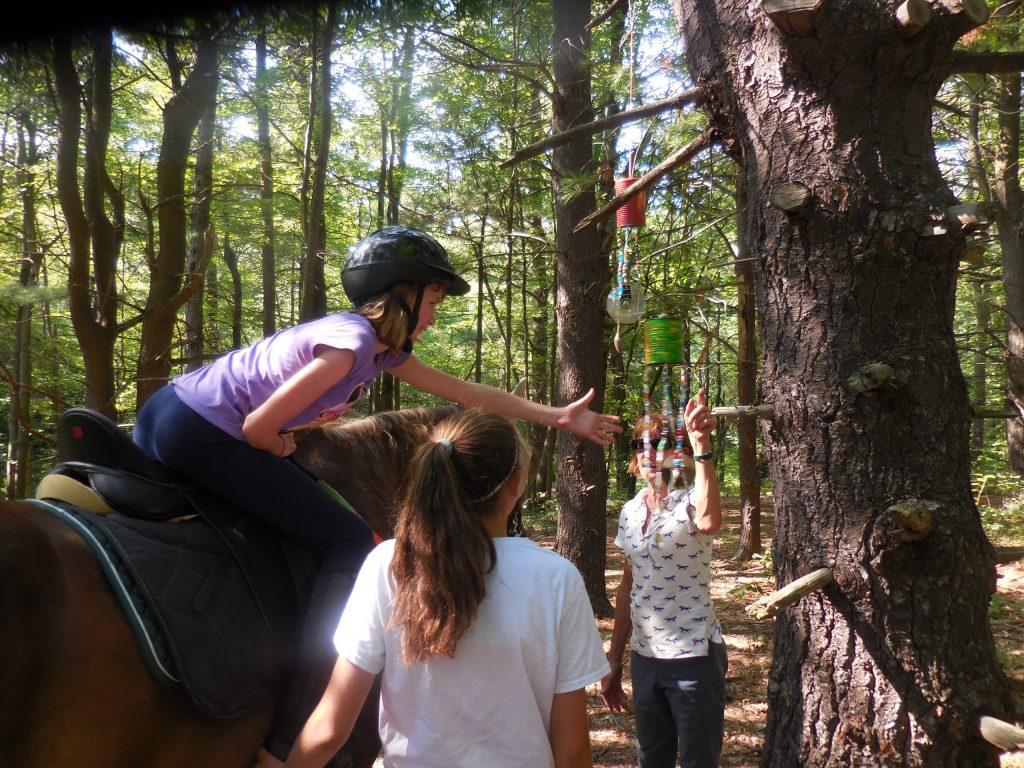 A girl on horseback with two women nearby reaches for an object hanging from a tree at High & Mighty's sensory trail course