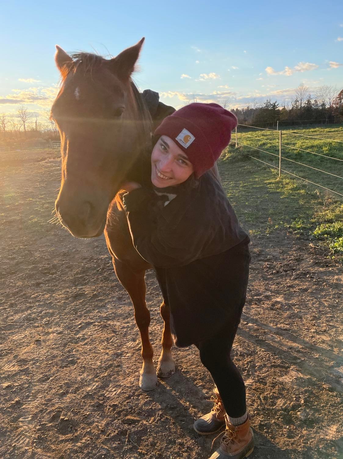 A woman wearing a red knit cap and hugging a brown horse's neck and smiling outdoors