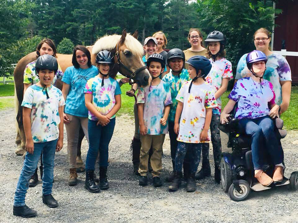 A row of kids smiling and wearing tie dye shirts with a brown horse with a white mane outside with green trees in the background