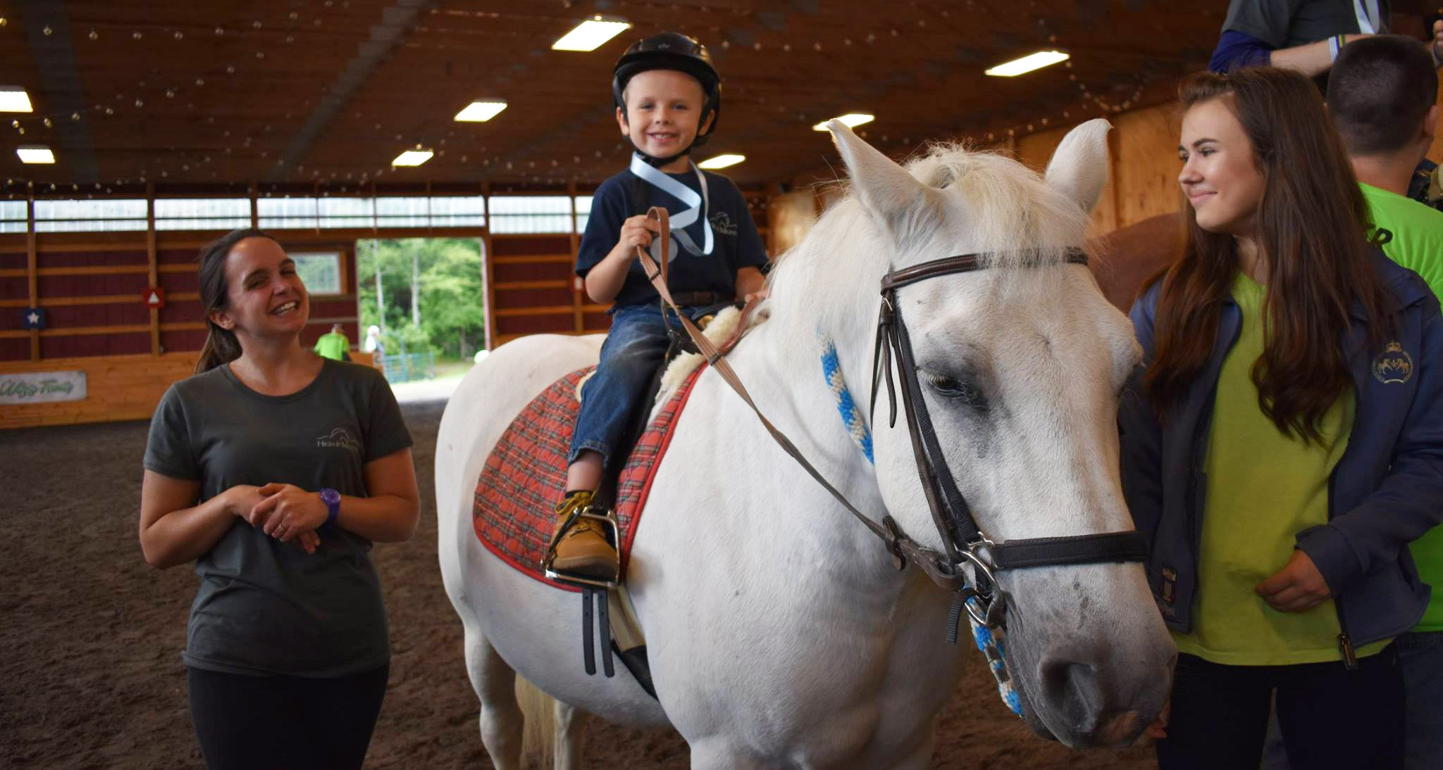 A young boy smiling on the back of a white pony with a woman smiling on the left and a girl on the right in the indoor ring at High & Mighty farm