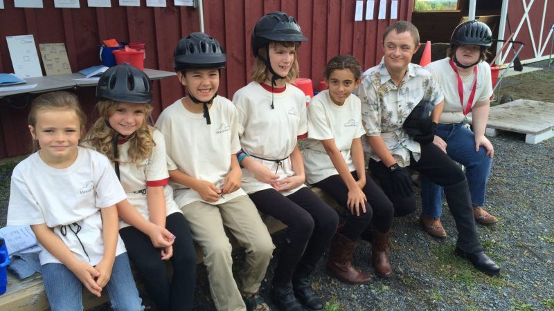 A row of kids sitting on a bench wearing hlemets in front of a red barn in High & Mighty's summer Horseplay camp