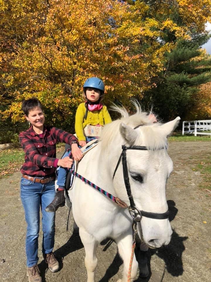 A woman on the left side of a white horse holding the reins while a girl in a yellow shirt rides and autumn leaves in the background