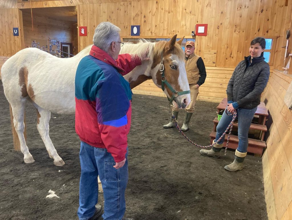 A man petting the neck of a brown and white paint horse while two adults stand nearby in the indoor ring in High & Mighty's veterans program