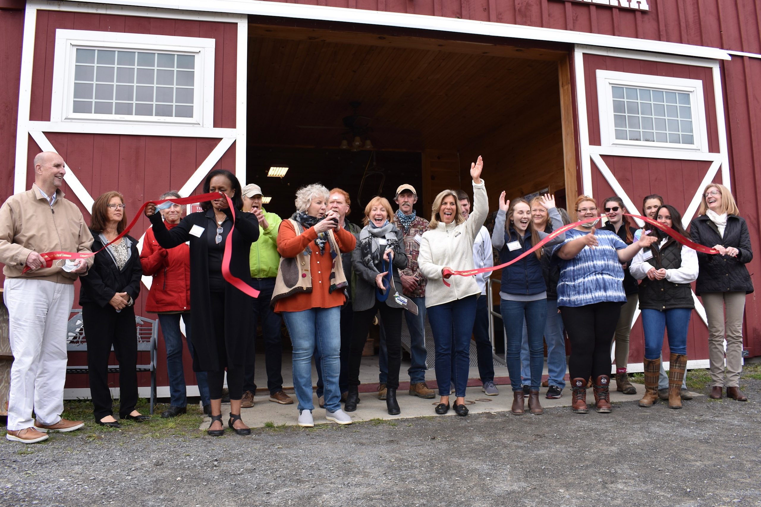 A group of people smiling and celebrating outside a red barn in the ribbon cutting ceremony at High & Mighty