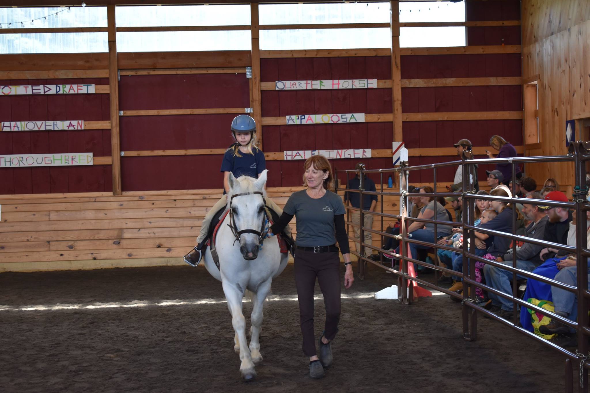 A young girl riding a white pony with a woman walking alongside to the right in the arena at High & Mighty